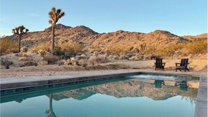 Ein Swimmingpool mit klarem Wasser, das die umliegende Landschaft spiegelt, darunter eine Wüste mit spärlicher Vegetation und ein Berg im Hintergrund. In der Nähe des Pools stehen zwei Holzstühle.