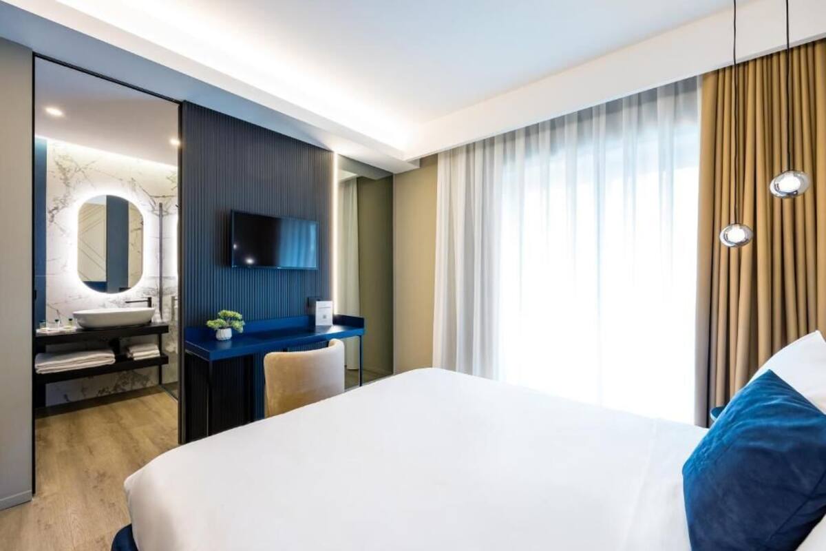 A modern hotel room with a large bed, a desk with a flat-screen TV, a vanity with a mirror and sink, and a view of the outdoors through large windows.