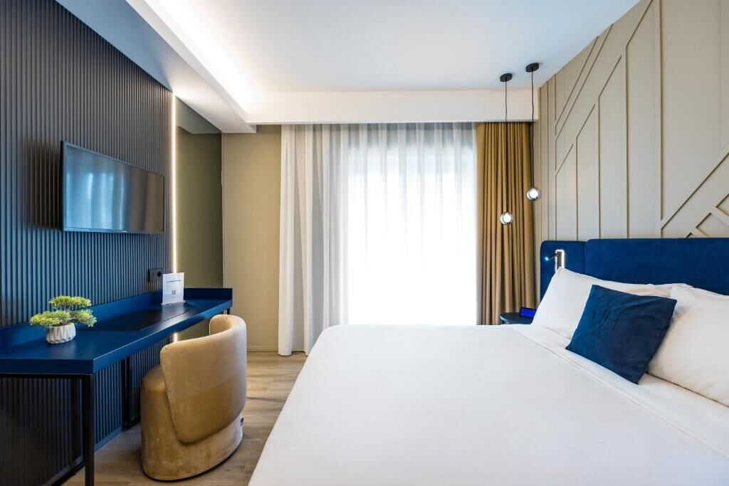 Une chambre d’hôtel moderne dotée d’un grand lit, d’un bureau avec un téléviseur à écran plat, d’un fauteuil confortable et offrant une vue sur l’extérieur grâce à de grandes fenêtres.