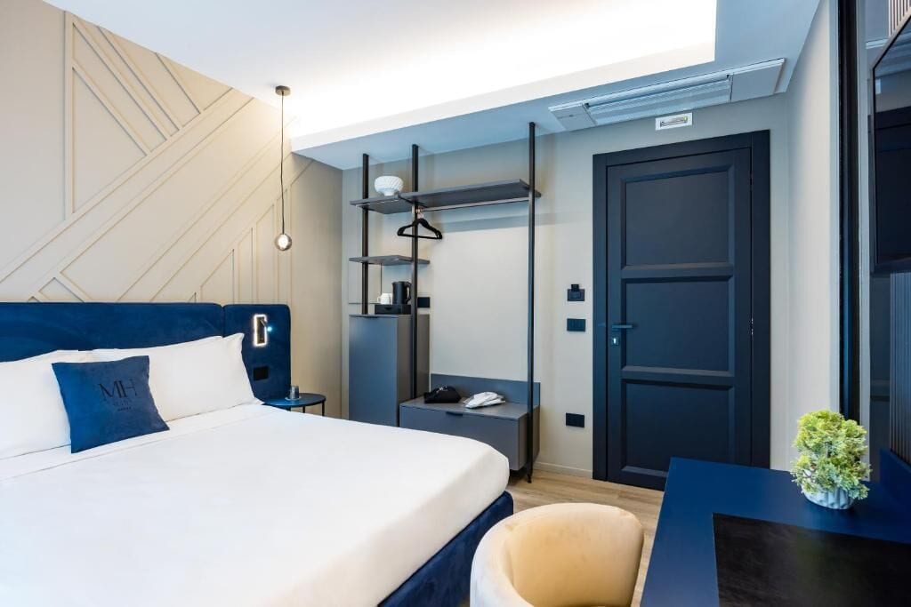 A modern hotel room with a neatly made bed, a bedside table with a lamp, a wardrobe, and a small desk with a chair.