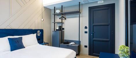 A modern hotel room with a neatly made bed, a bedside table with a lamp, a wardrobe, and a small desk with a chair.