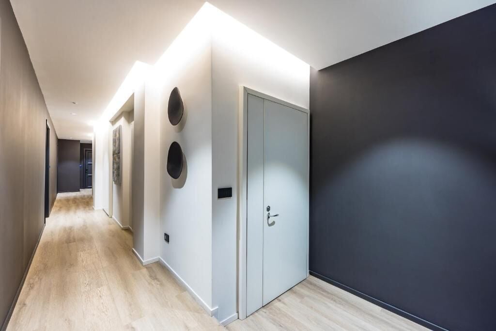 A modern hallway with a light wooden floor, a dark wall on the right, and a light-colored door with a handle.