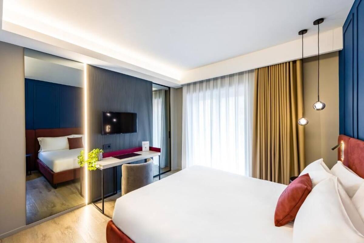 A modern hotel room with a large bed, a desk with a chair, a flat-screen TV, and a large window with curtains.