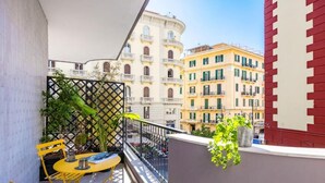 A balcony with yellow chairs and a table, a black metal fence, and a view of buildings in the background. - MH SUITES PLEBISCITO (Naples)
