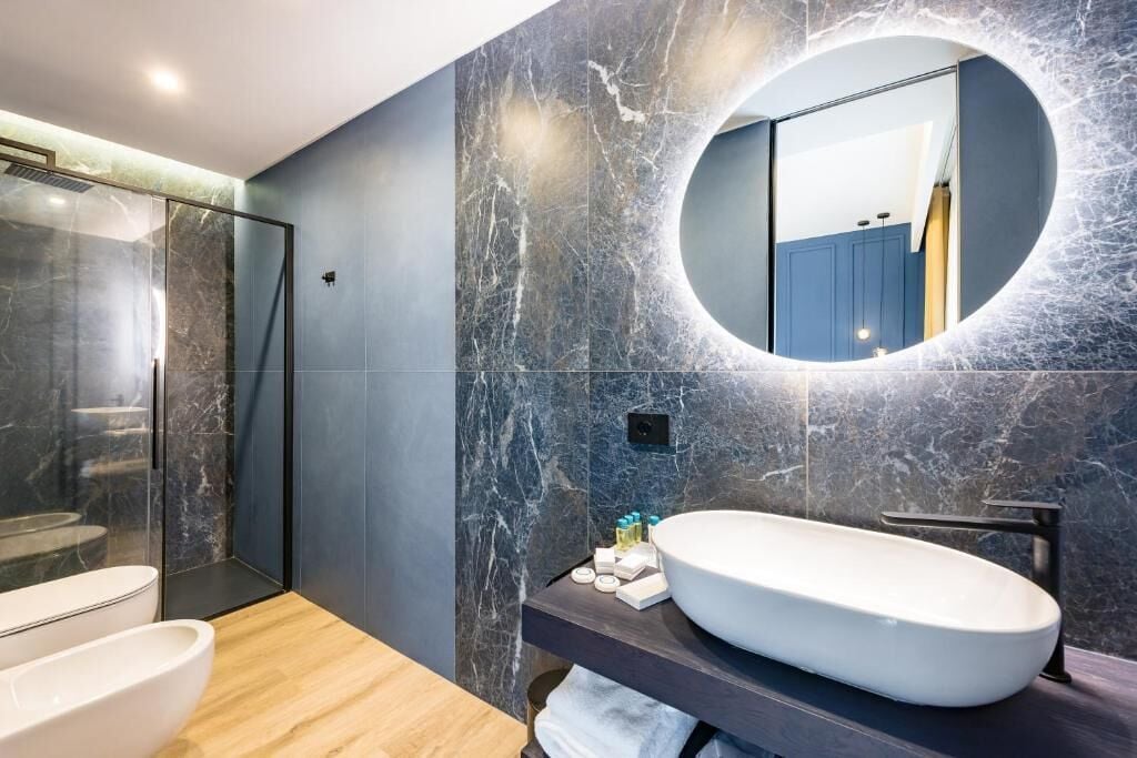 Un baño moderno con un lavamanos ovalado grande, un espejo redondo y una ducha con mampara de vidrio.