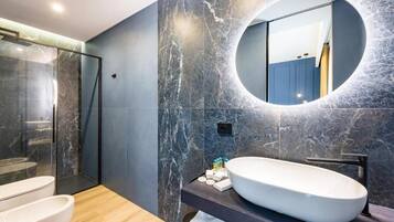 Un baño moderno con un lavamanos ovalado grande, un espejo redondo y una ducha con mampara de vidrio.