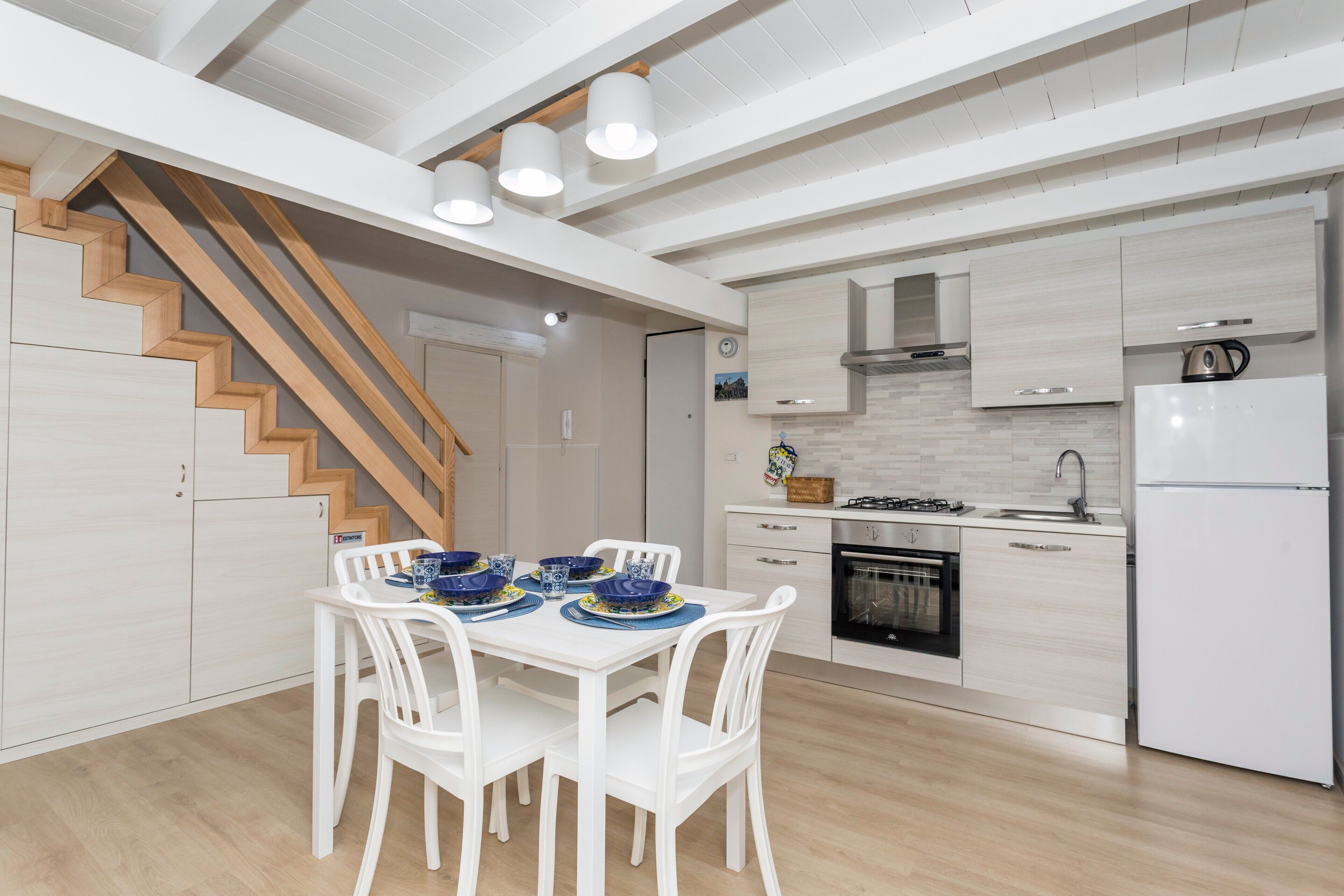 Une cuisine moderne avec des meubles blancs, une cuisinière encastrée et un réfrigérateur. On y trouve un coin repas avec une table et des chaises. Un escalier en bois est visible au fond.