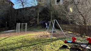 Un terrain de jeux avec des balançoires et un toboggan, entouré d’un mur de pierre et d’arbres.