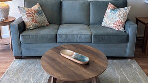 A living room with a grey sofa, two patterned pillows, a wooden coffee table, and a large abstract painting on the wall. - New Full Remodel 2nd Floor Oceanfront Condo (Kihei)