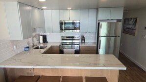 A modern kitchen with stainless steel appliances, a marble countertop, and wooden stools. - New Full Remodel 2nd Floor Oceanfront Condo (Kihei)