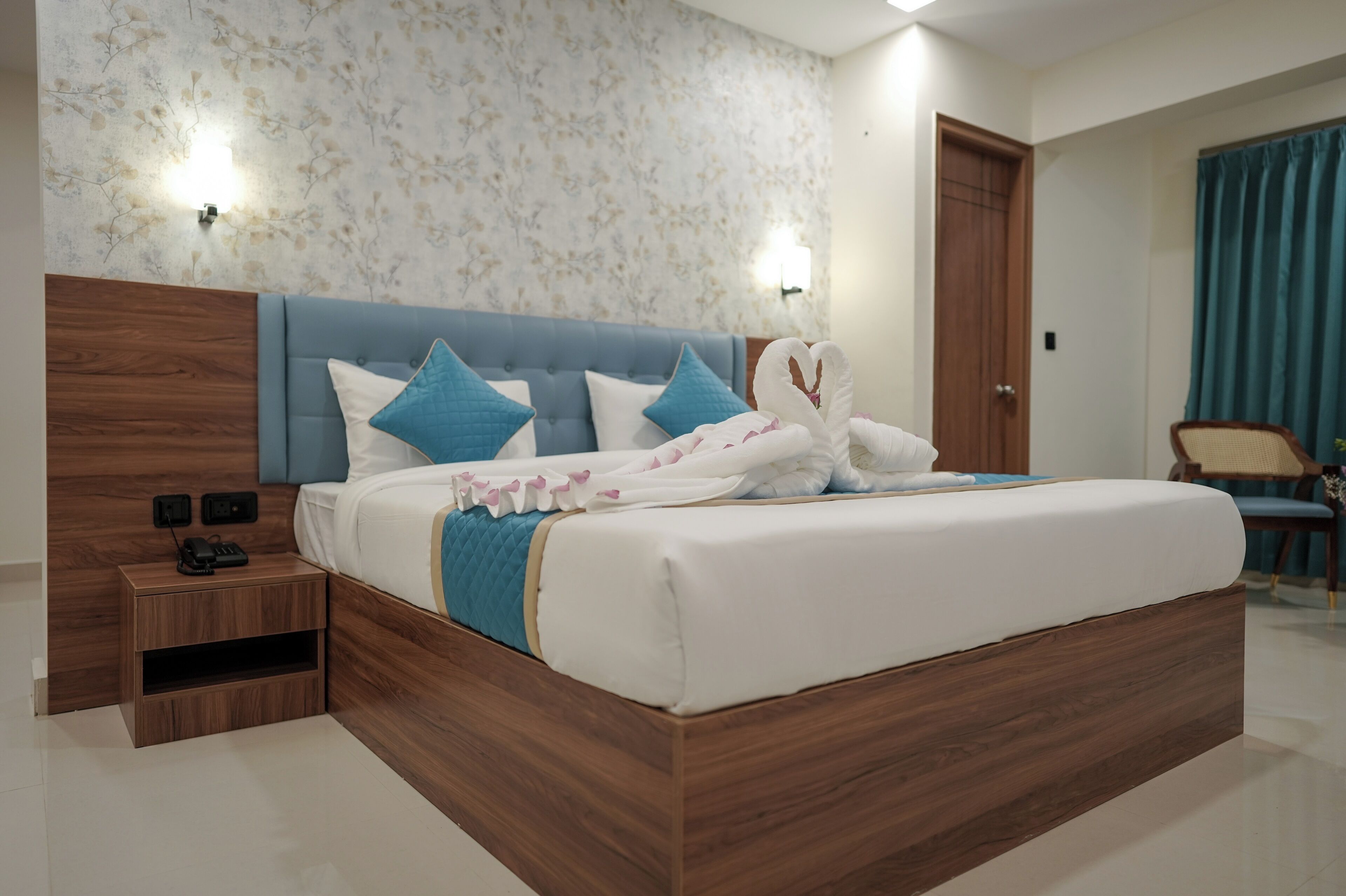 Een moderne slaapkamer met een houten hoofdeinde, een bed met witte lakens en blauwe kussens, een nachtkastje met een telefoon, en bloemenbehang.