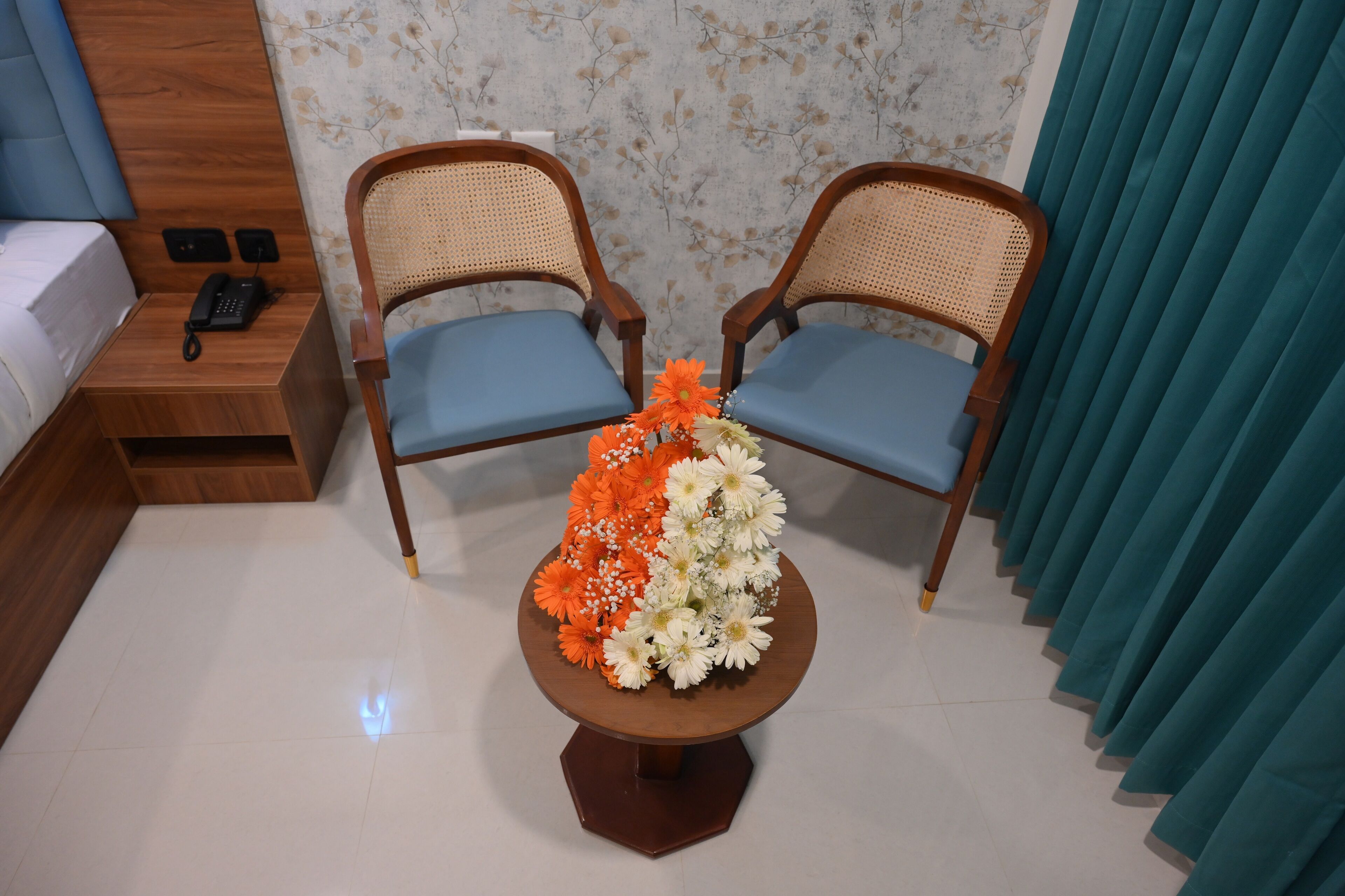 A hotel room with two wooden chairs with blue cushions, a small wooden table with a floral centerpiece, a bedside table with a phone, and a curtain.