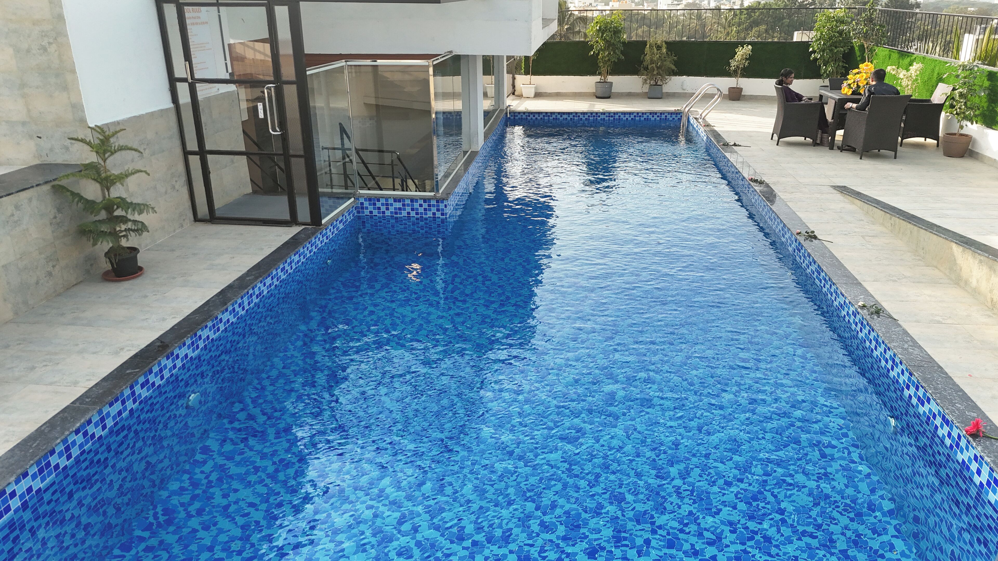 A rectangular swimming pool with blue mosaic tiles, surrounded by a patio area with outdoor furniture and potted plants.