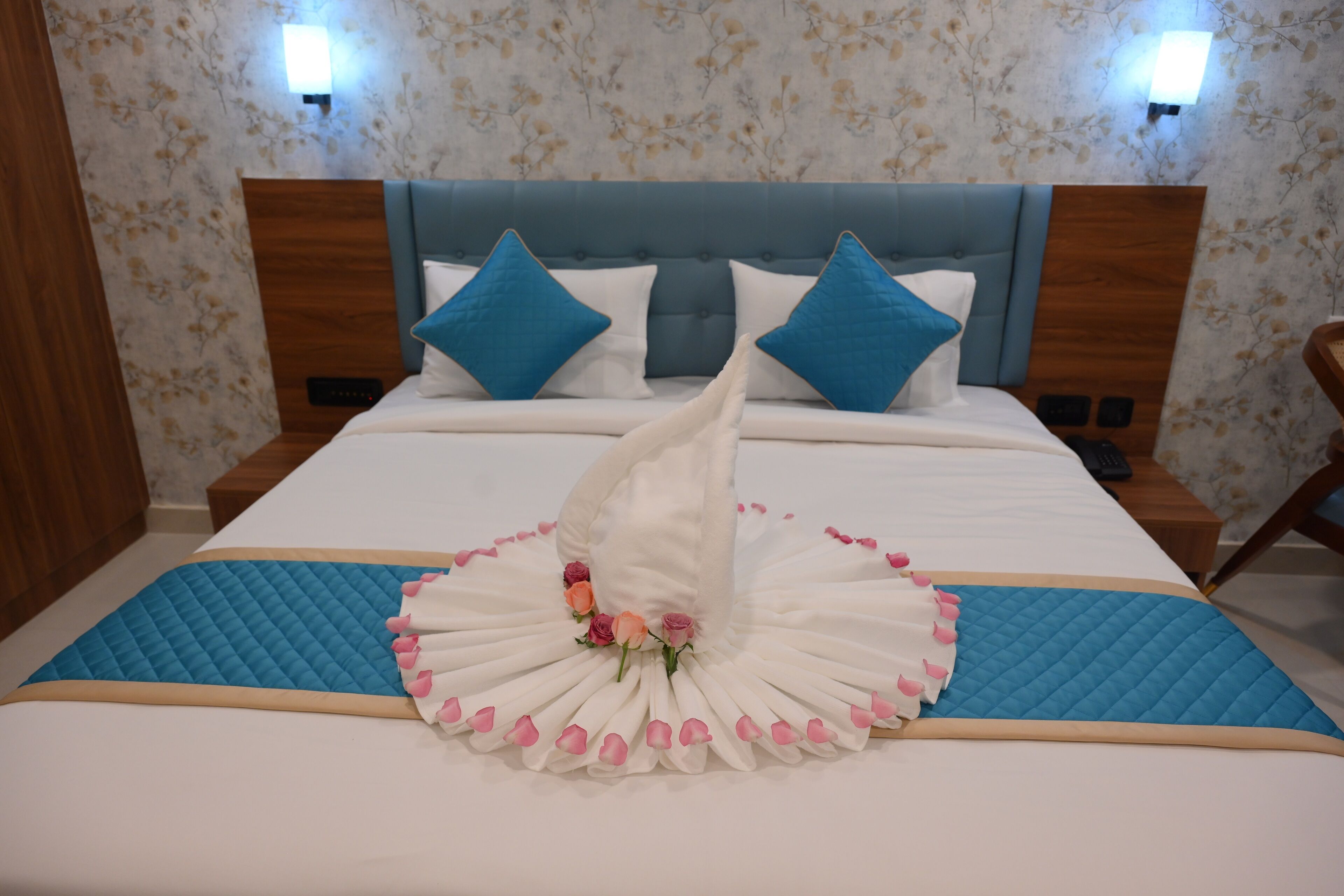 A neatly made bed with a decorative towel and flowers, a blue quilted bedspread, and two blue pillows on a wooden headboard.