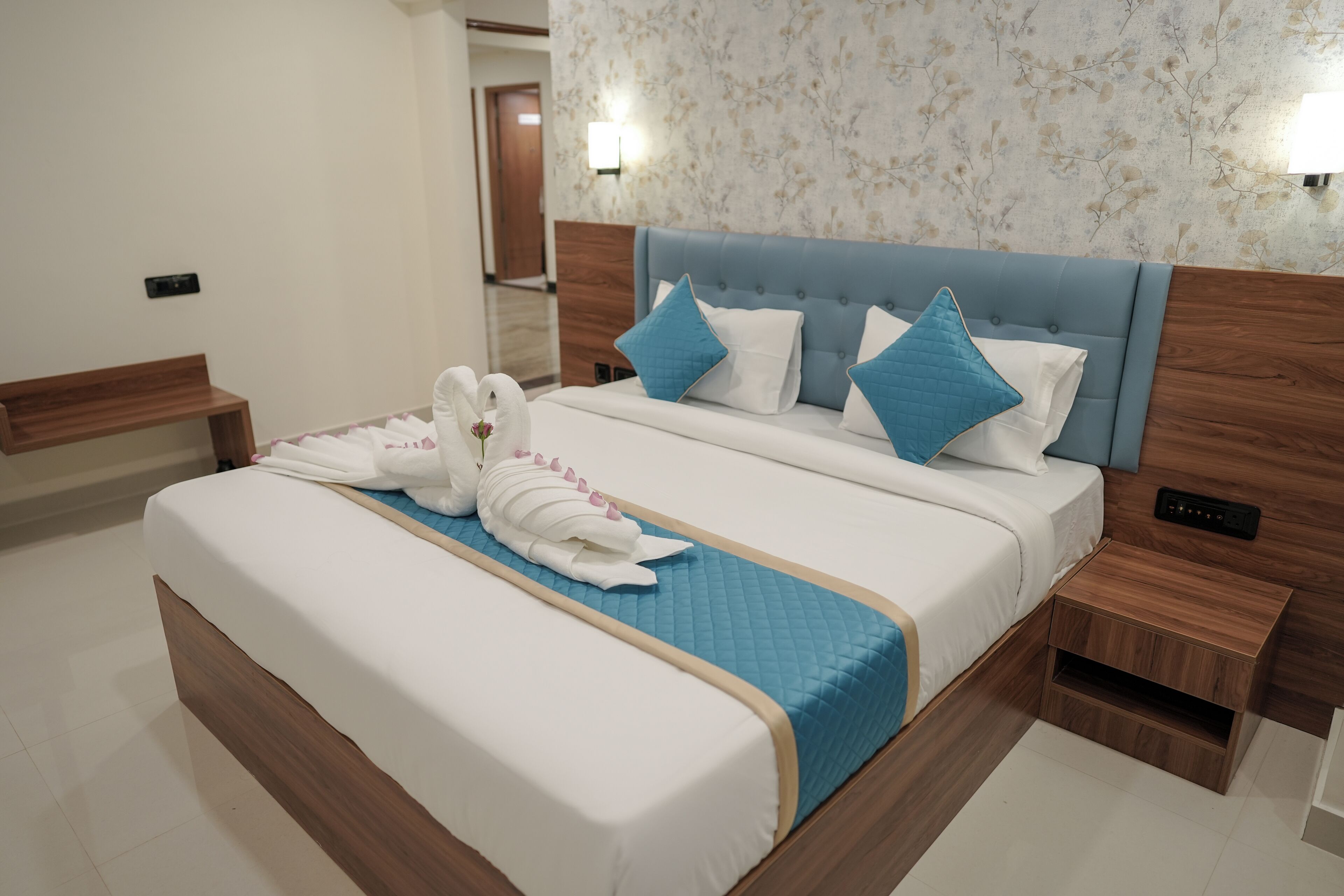 A neatly made bed with white and blue bedding, two towels, and decorative pillows. There is a wooden nightstand with a lamp beside the bed.