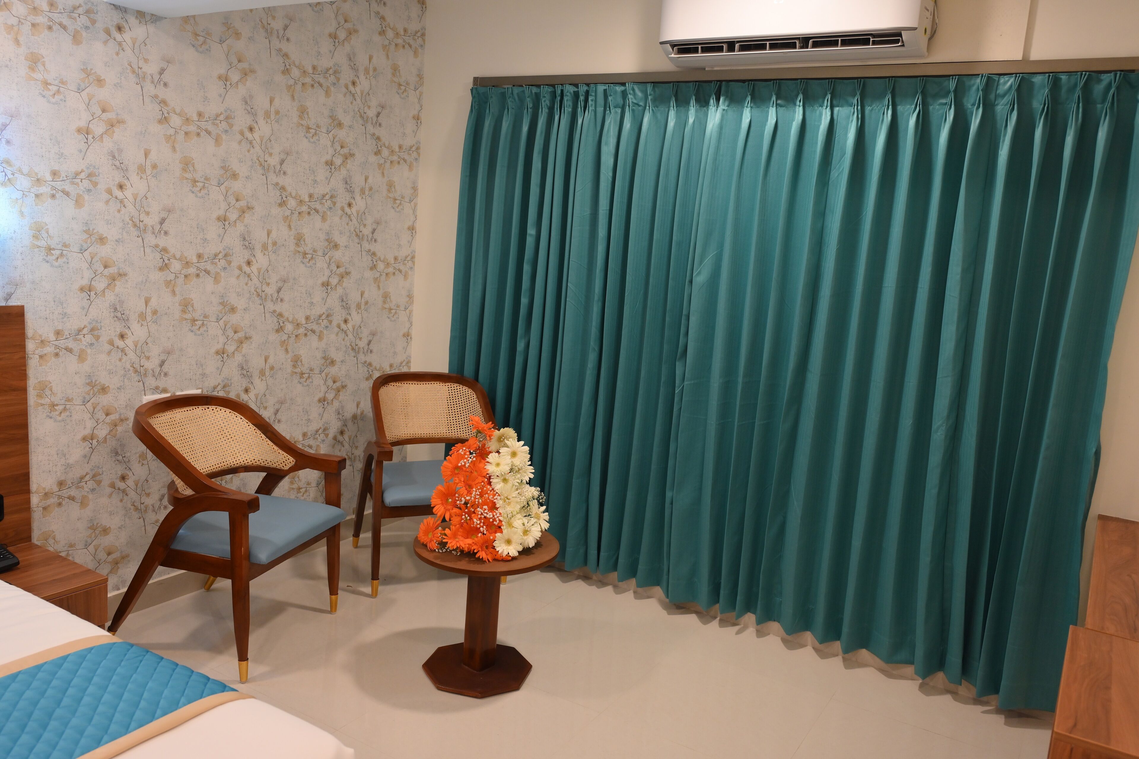A hotel room with two wooden chairs, a small table with a floral arrangement, a bed with a blue quilt, and teal curtains.