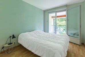 Un dormitorio con cama, escritorio con lámpara, ventana y puerta corrediza de vidrio a un balcón.
