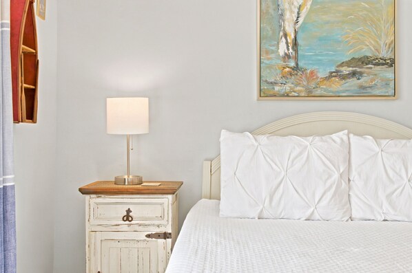 Um quarto com uma cama, uma mesinha de cabeceira com abajur, cômoda de madeira e um quadro na parede.
