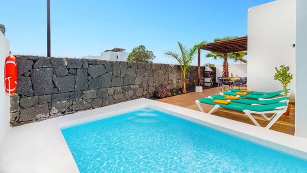 A swimming pool with lounge chairs, a stone wall, and a pergola. - Villa Sapri by Villa Plus (Playa Blanca)