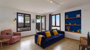 A living room with a blue sofa, a pink armchair, and a small table with a plant. There are large windows, a blue shelf with decorative items, and a TV stand with a remote control. - Villa Sapri by Villa Plus (Playa Blanca)
