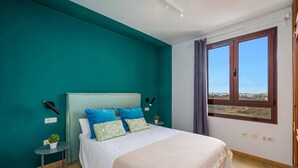 A bedroom with a bed, a nightstand, a potted plant, a window with a view, and teal walls. - Villa Sapri by Villa Plus (Playa Blanca)