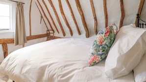 Ein Bett mit weißer Bettwäsche und einem blumigen Kopfkissen, Holzbalken an der Wand und ein Fenster mit Vorhängen.