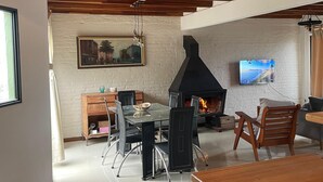 A modern living room with a fireplace, wooden ceiling beams, a dining table, and a TV mounted on the wall.