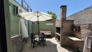 A patio with a table and chairs under a large umbrella, a brick oven, and a green wall with an air conditioning unit.