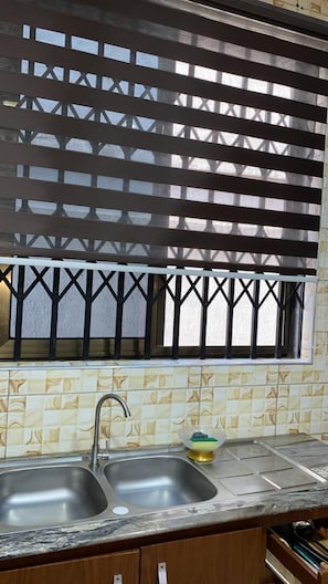 A kitchen with a double sink, a window with a metal grill, and a patterned backsplash. - NickJaak Courtyard Rooms (Accra)