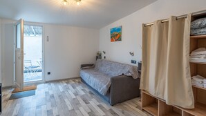 Een kamer met een bank, houten vloer, een deur naar buiten, een plank met opgevouwen kleding en een schilderij aan de muur.