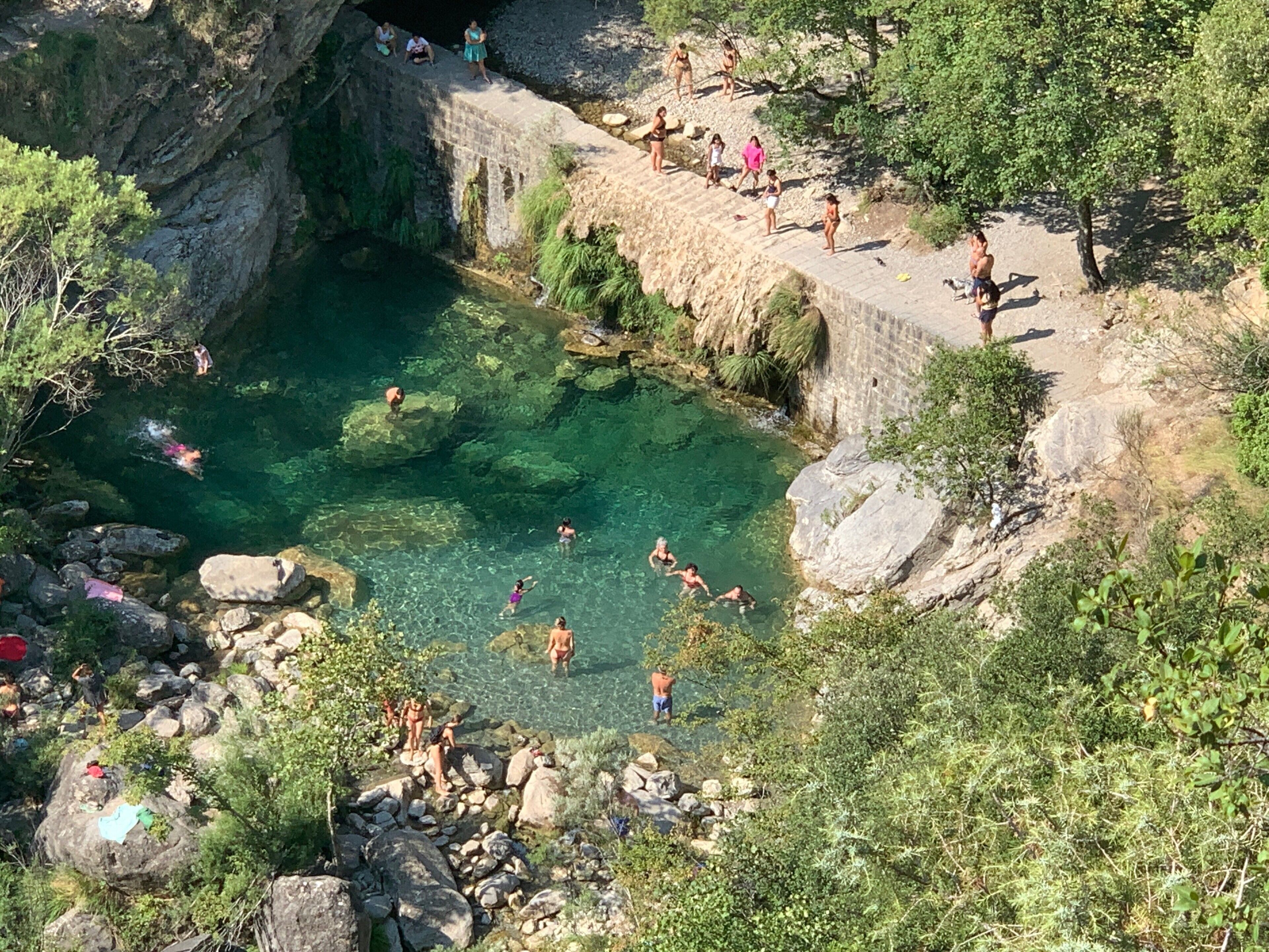 Ein natürlicher Pool, umgeben von felsigem Gelände und viel Grün, mit darin schwimmenden und sich aufhältenden Personen.