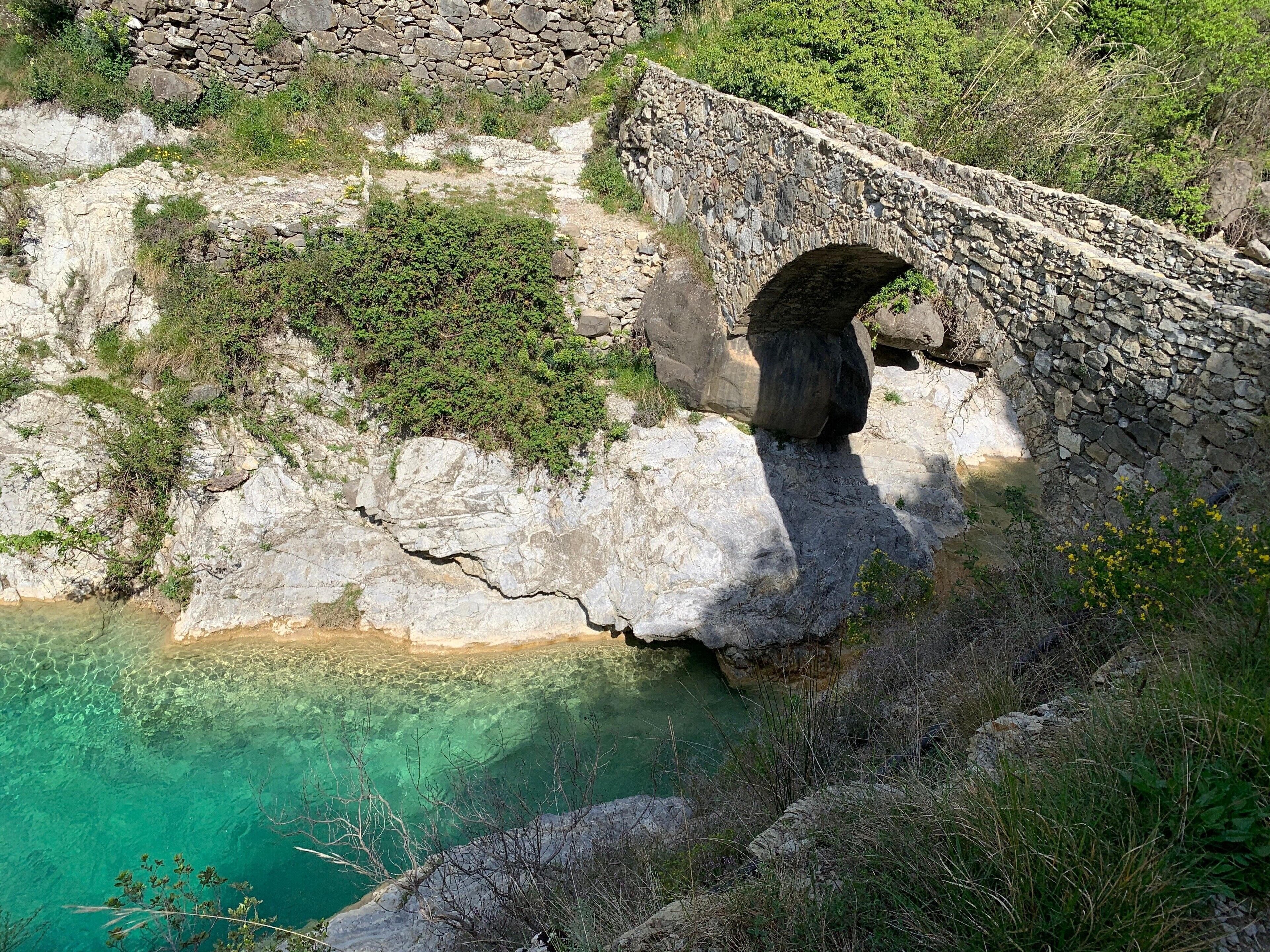 A stone bridge over a clear blue pool, surrounded by rocky terrain and greenery.