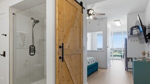 Un interno moderno con cabina doccia in vetro, una porta scorrevole in legno, un letto con testiera blu e vista sull'esterno.