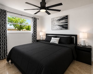 A bedroom with a black bedspread, a ceiling fan, a nightstand with a lamp, and a window with patterned curtains. - CASA KAMPA offers you a heated pool with Jacuzzi at no additional cost. (Tampa)