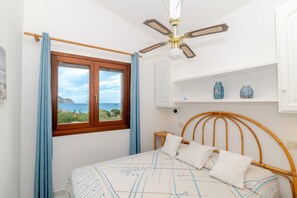 Ein Schlafzimmer mit Bett, Deckenventilator, Fenster mit Vorhängen und Blick aufs Meer.