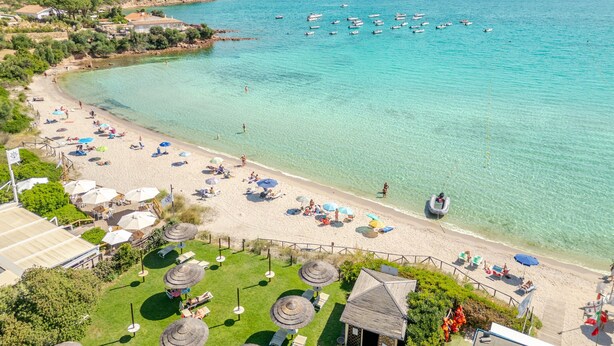 Ein Strand mit klarem türkisblauem Wasser, ein Berg im Hintergrund und mehrere Boote.