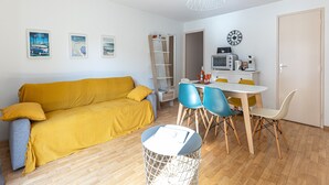 A living room with a yellow sofa, a dining table with chairs, a white lamp, and a small white trash bin.