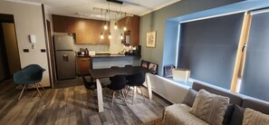 A modern kitchen with wooden cabinets, a stainless steel refrigerator, and a dining table with chairs. There is a blue chair and a sofa with a knitted throw.