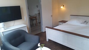 Une chambre d’hôtel comprenant un lit, une télévision, une chaise, une petite table avec un vase et une porte donnant sur un coin repas.