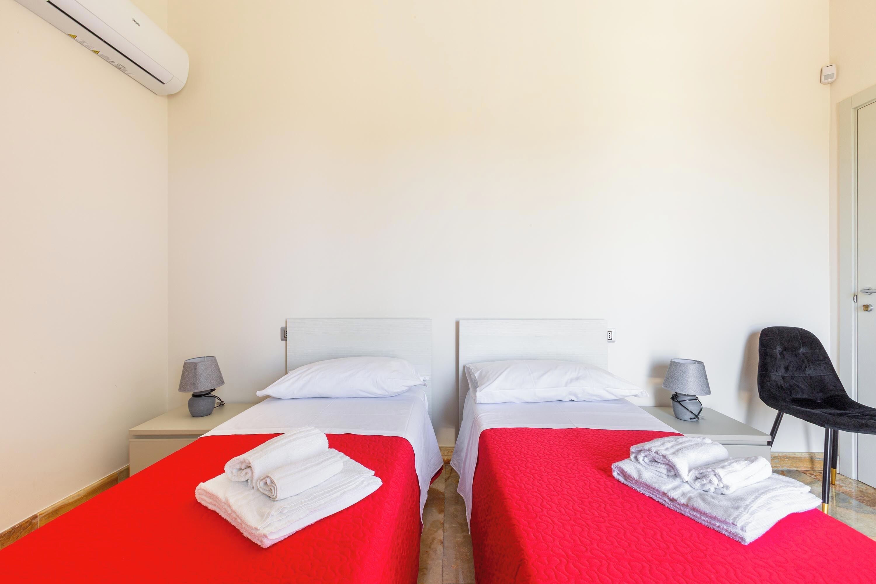 A hotel room with two beds, each with a red bedspread and white pillows, and a black chair.