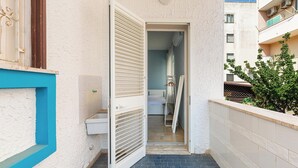 Un couloir étroit avec une fenêtre à cadre bleu, une porte blanche dotée de stores horizontaux et un sol carrelé.