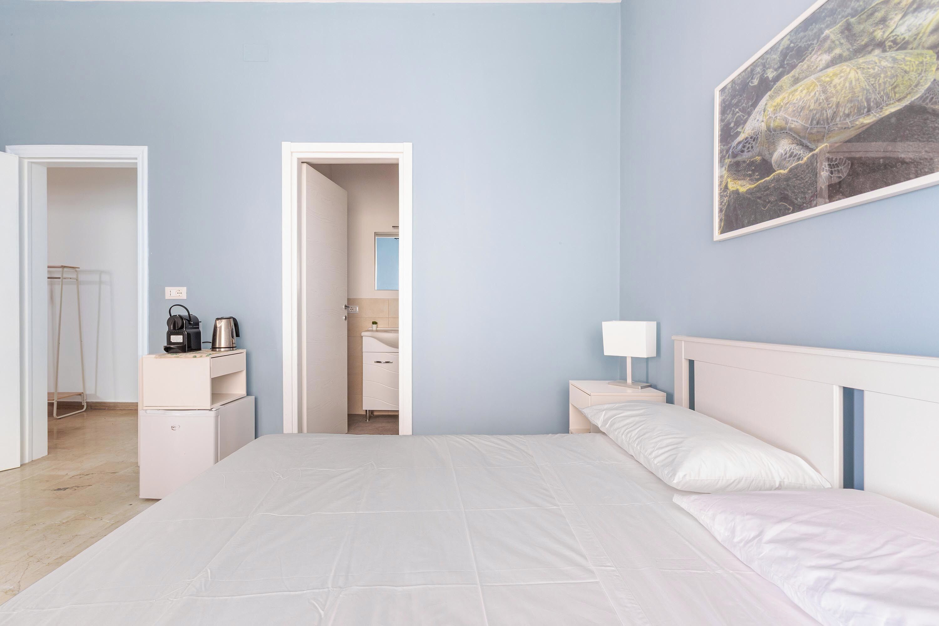 Ein Schlafzimmer mit einem großen Bett, einem Nachttisch, einer Kaffeemaschine und einem gerahmten Bild an der Wand.