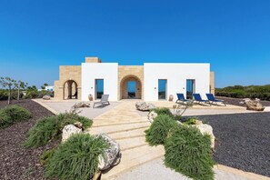 Un bâtiment moderne doté d’une arche centrale, de deux ailes latérales et d’une allée pavée menant à un espace aménagé avec des chaises bleues.
