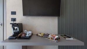Une chambre d’hôtel moderne équipée d’un téléviseur à écran plat fixé au mur, d’une machine à café, de quelques en-cas, d’un plateau avec une tasse et une soucoupe, et d’un livre posé sur la table.