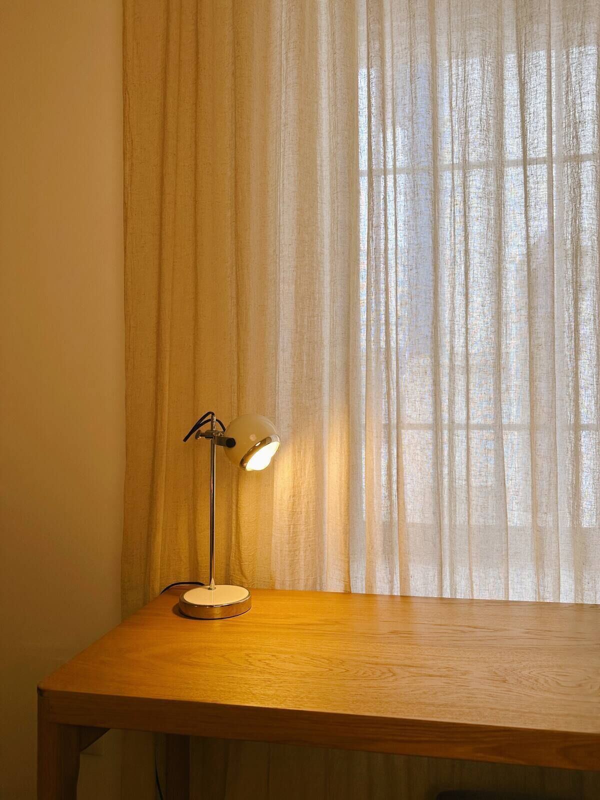 A desk lamp with a white shade and a metallic base, positioned on a wooden table in front of a sheer curtain.