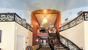Una majestuosa escalera con barandales ornamentados, un candelabro y un techo alto con un mural.