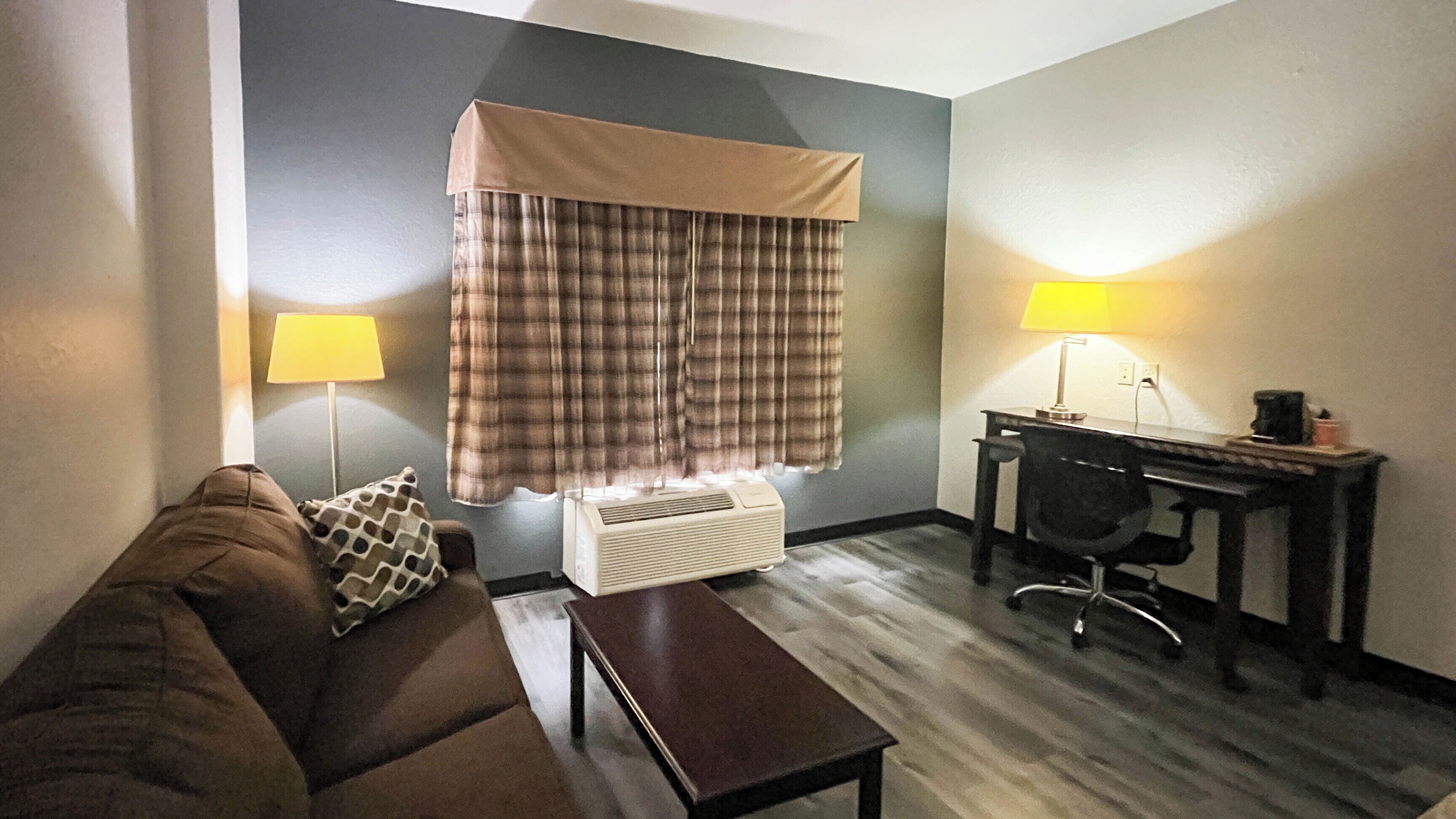Une chambre d’hôtel équipée d’un canapé, d’un bureau, d’une lampe, d’un climatiseur et d’une fenêtre avec des rideaux.