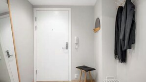 Ein Zimmer mit weißer Tür, einem kleinen Hocker, einem Kleiderständer mit Jacken und einem weißen Regal.