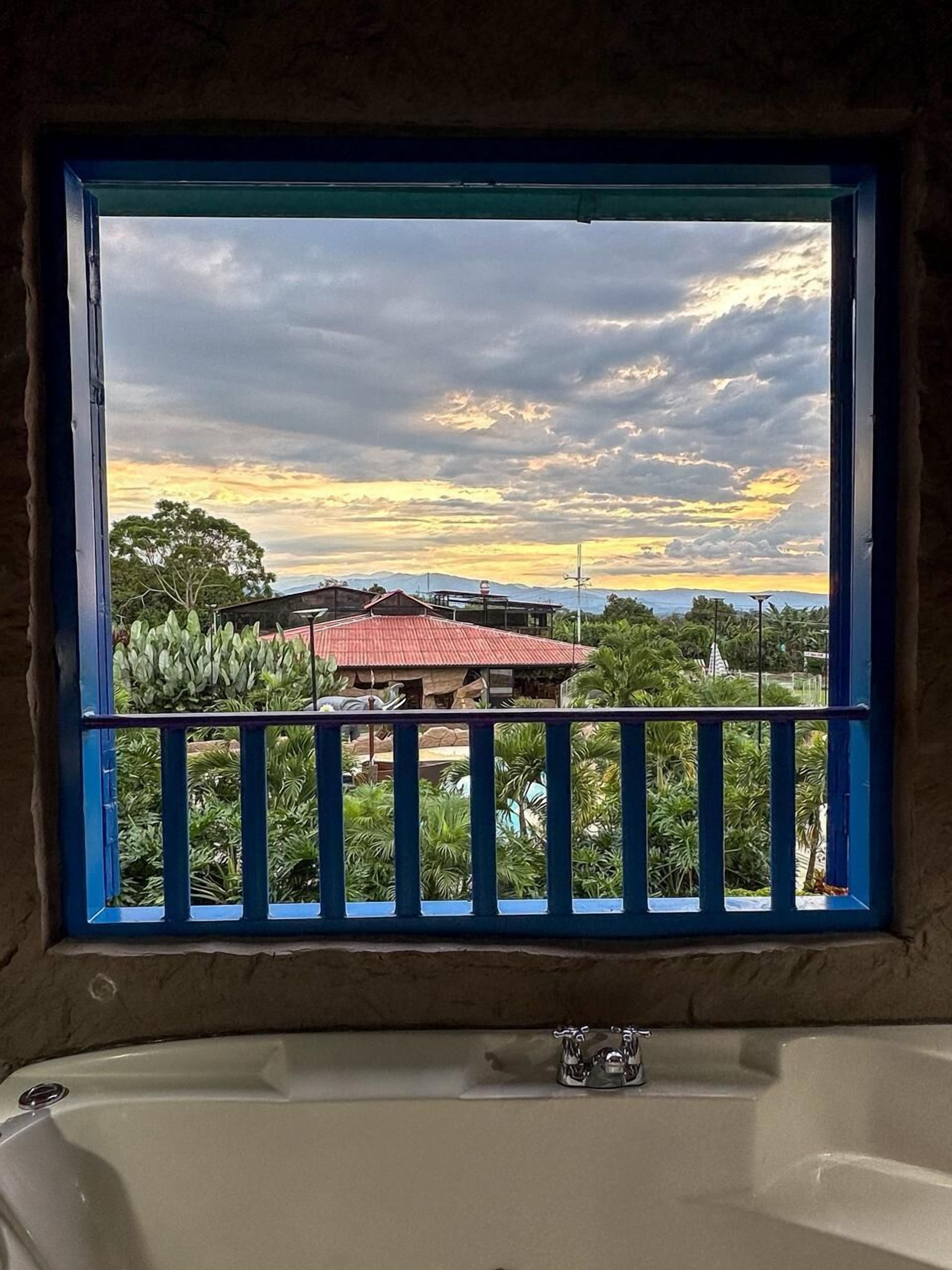 A bathroom with a bathtub, a window with a blue frame, and a view of a red-roofed building and greenery.