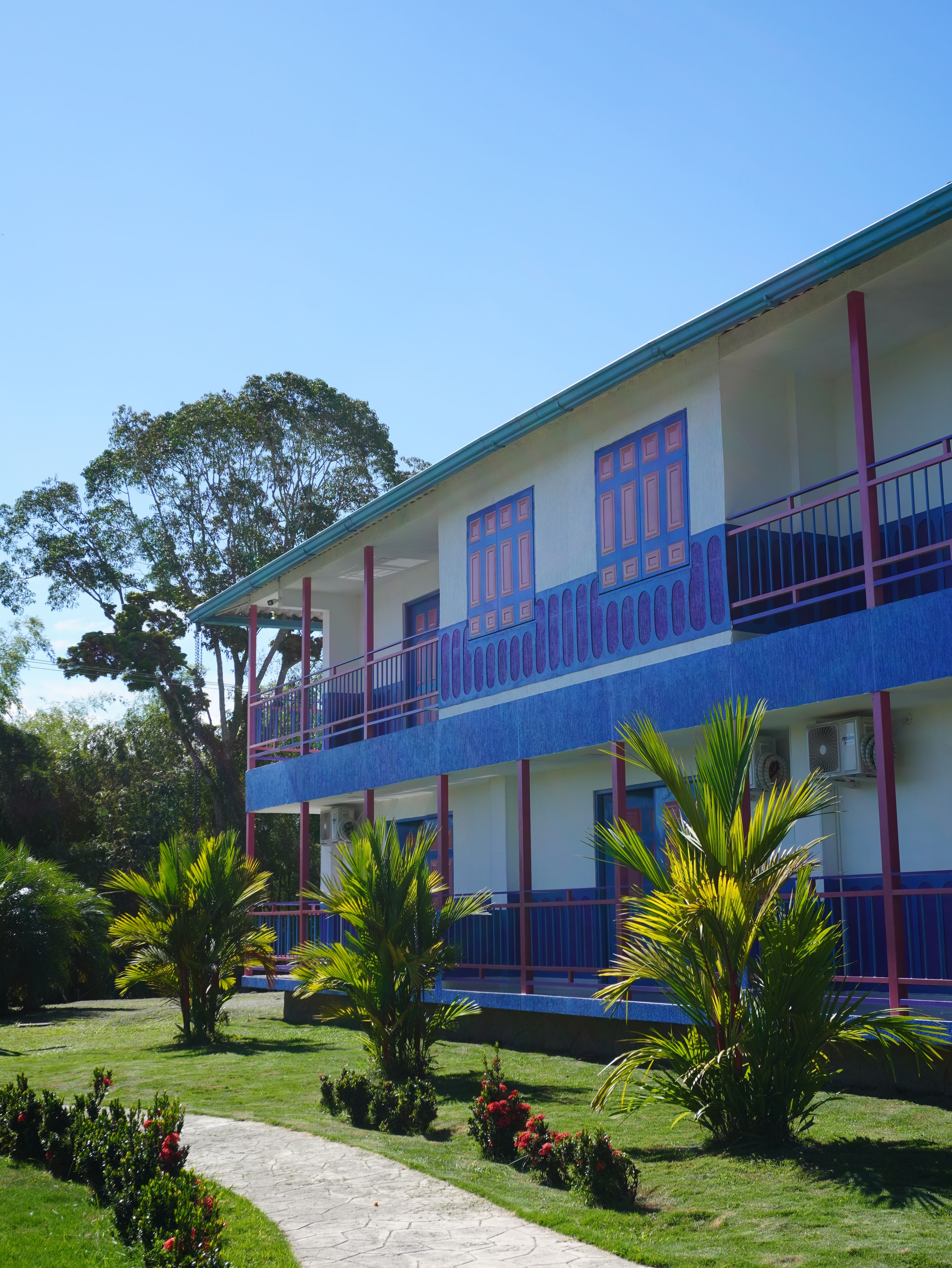 A two-story building with a balcony, surrounded by a well-maintained garden with palm trees and red flowers.